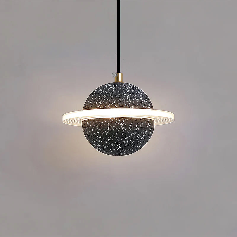 Sand black speckled Clérachic pendant with a brass cord top and a bright ring, lit against a grey wall.