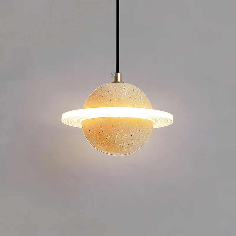 A Clérachic pendant with a sand yellow sphere, brass cord top and bright ring, glowing against a grey wall.
