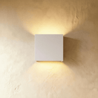 This Clérachic square wall lamp in matte white finish casts warm light up and down a plaster wall.