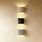 Three Clérachic square wall lamps in gold, black and silver finishes are stacked vertically on a wall.