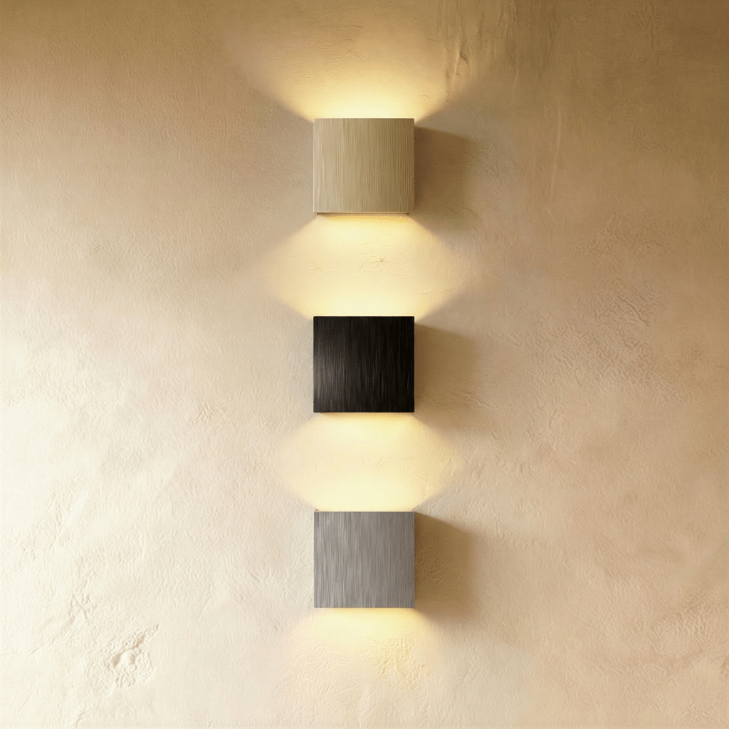 Three Clérachic square wall lamps in gold, black and silver finishes are stacked vertically on a wall.