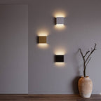 Three Clérachic square wall lamps in silver, gold and black light a gray wall beside a terracotta vase.