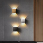 Three Clérachic square wall lamps in gold, silver and black glow on a paneled wall above a dark sofa.