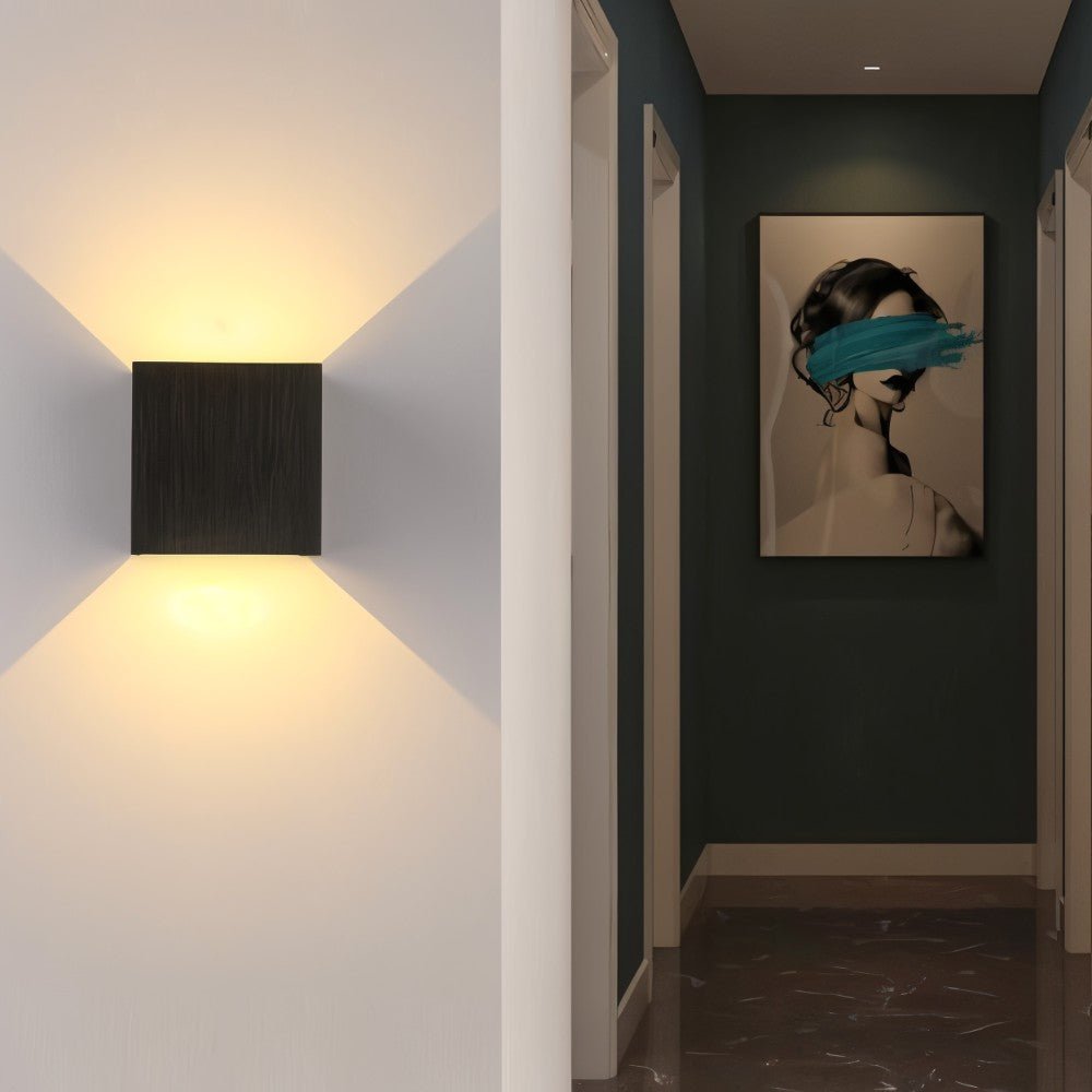 A Clérachic square wall lamp in a dark brushed finish lights a white hallway wall near framed art.