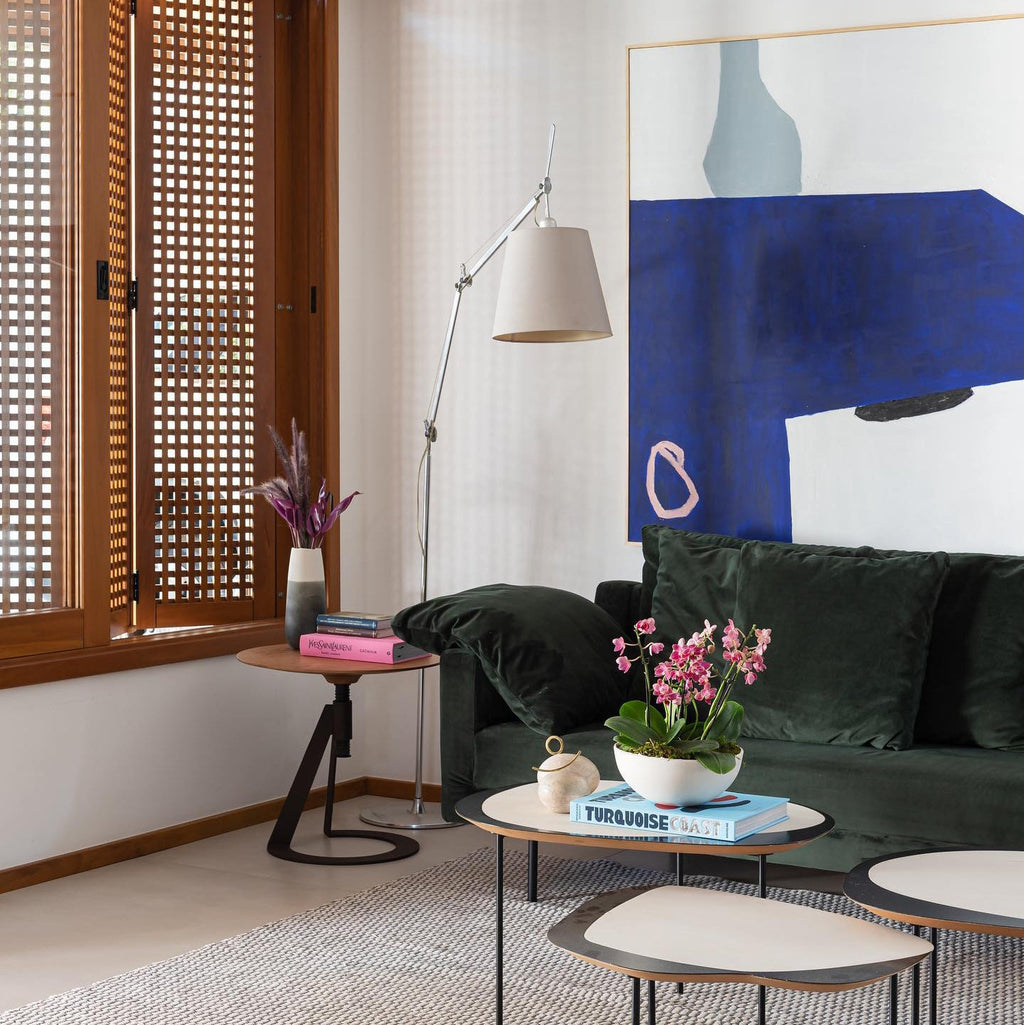 Silver-armed Clérachic floor lamp behind a dark green sofa, beside a large blue abstract painting and a pair of low tables.