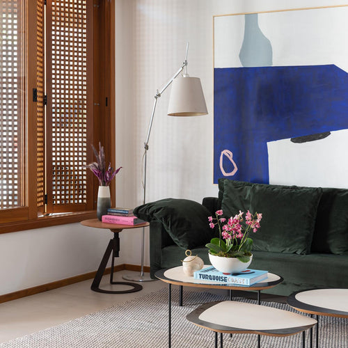 Silver-armed Clérachic floor lamp behind a dark green sofa, beside a large blue abstract painting and a pair of low tables.