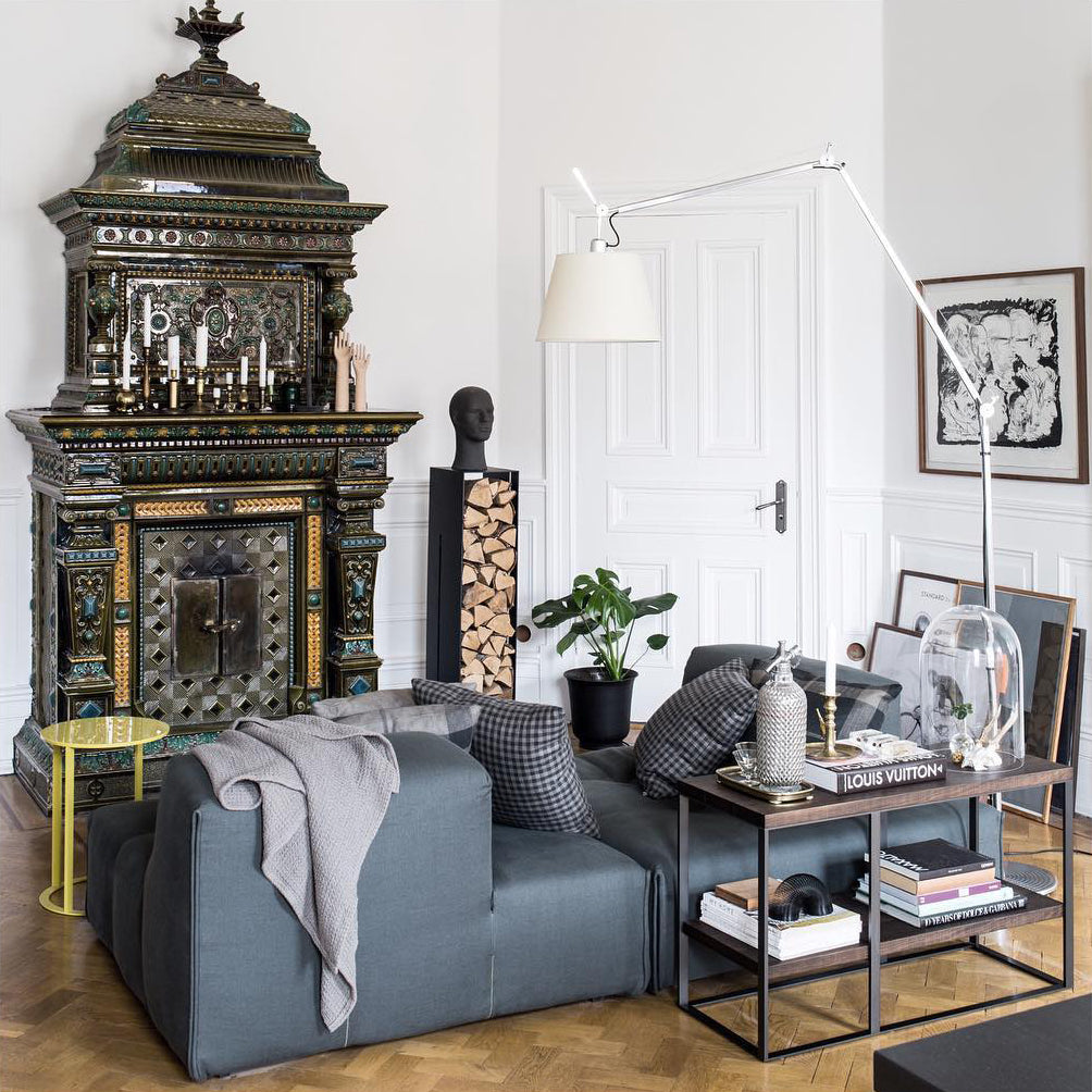Clérachic arm floor lamp over a grey sofa in a room with a tiled stove, a white panelled door and framed artwork.