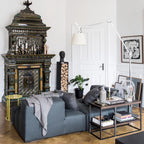 Clérachic arm floor lamp over a grey sofa in a room with a tiled stove, a white panelled door and framed artwork.