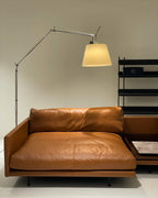 Lit Clérachic floor lamp with a warm cream shade, arched over a tan leather chaise in a pale room.