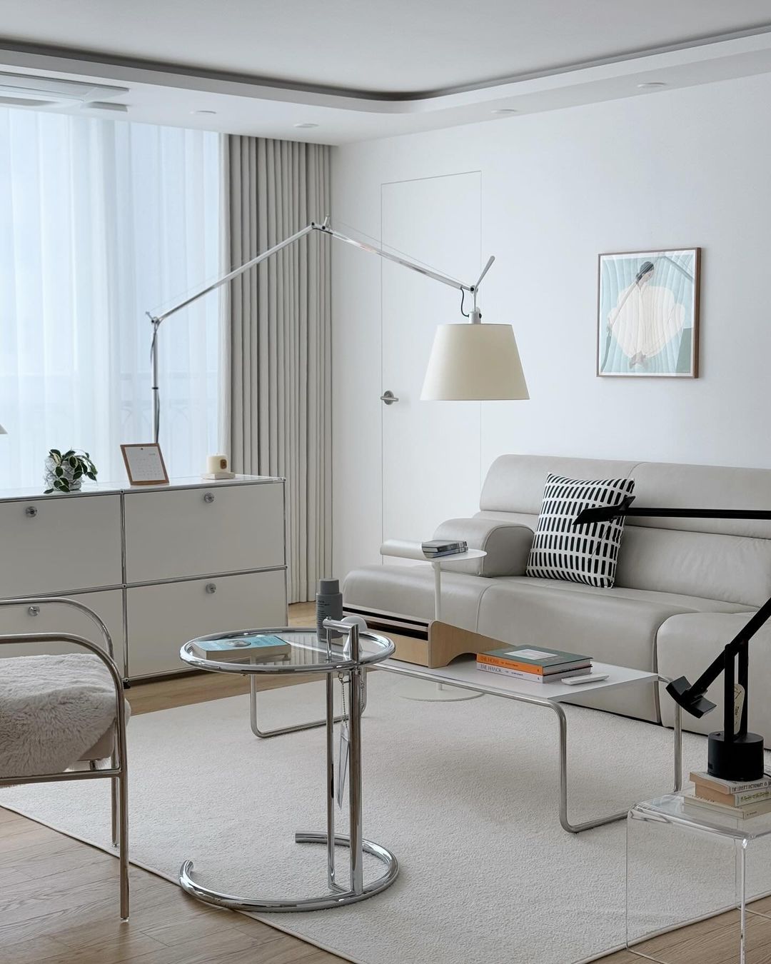 Clérachic arc floor lamp in a white living room with a pale sofa, a glass side table and low white cabinets.