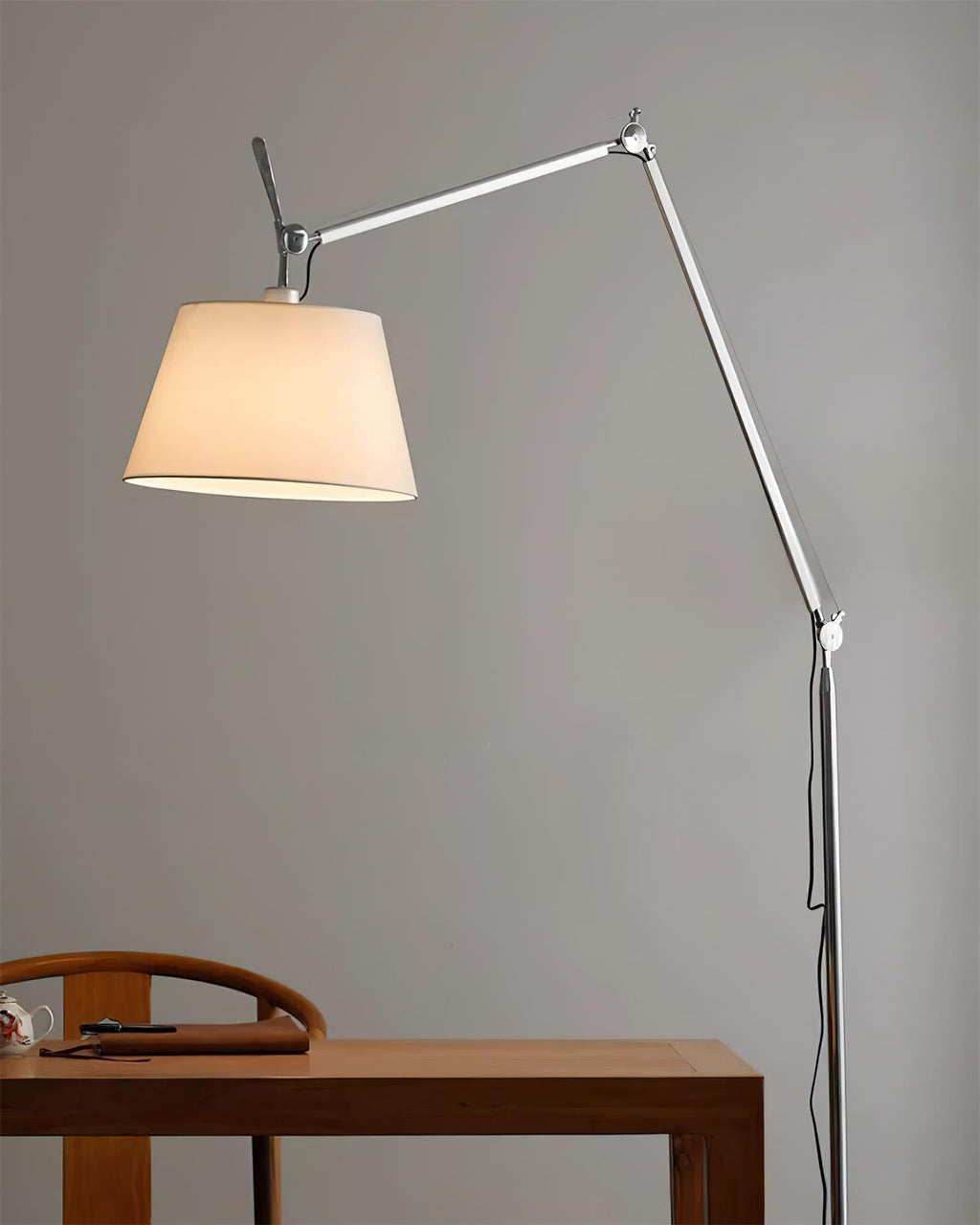 Clérachic floor lamp with a lit cream shade against a grey wall, above a wooden table and chair.