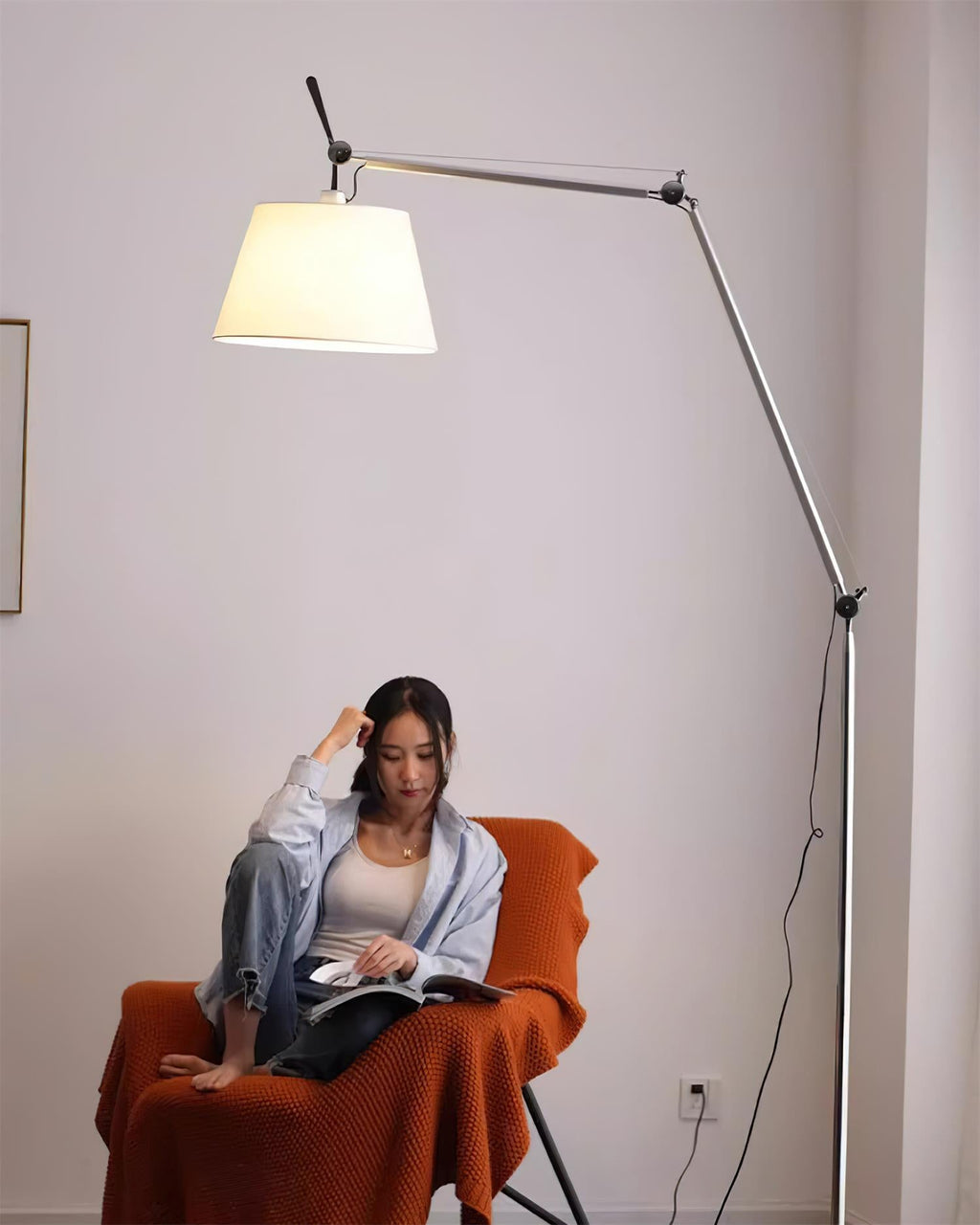 A woman reads in an orange armchair under the lit shade of this Clérachic floor lamp, its cord trailing down the pole.