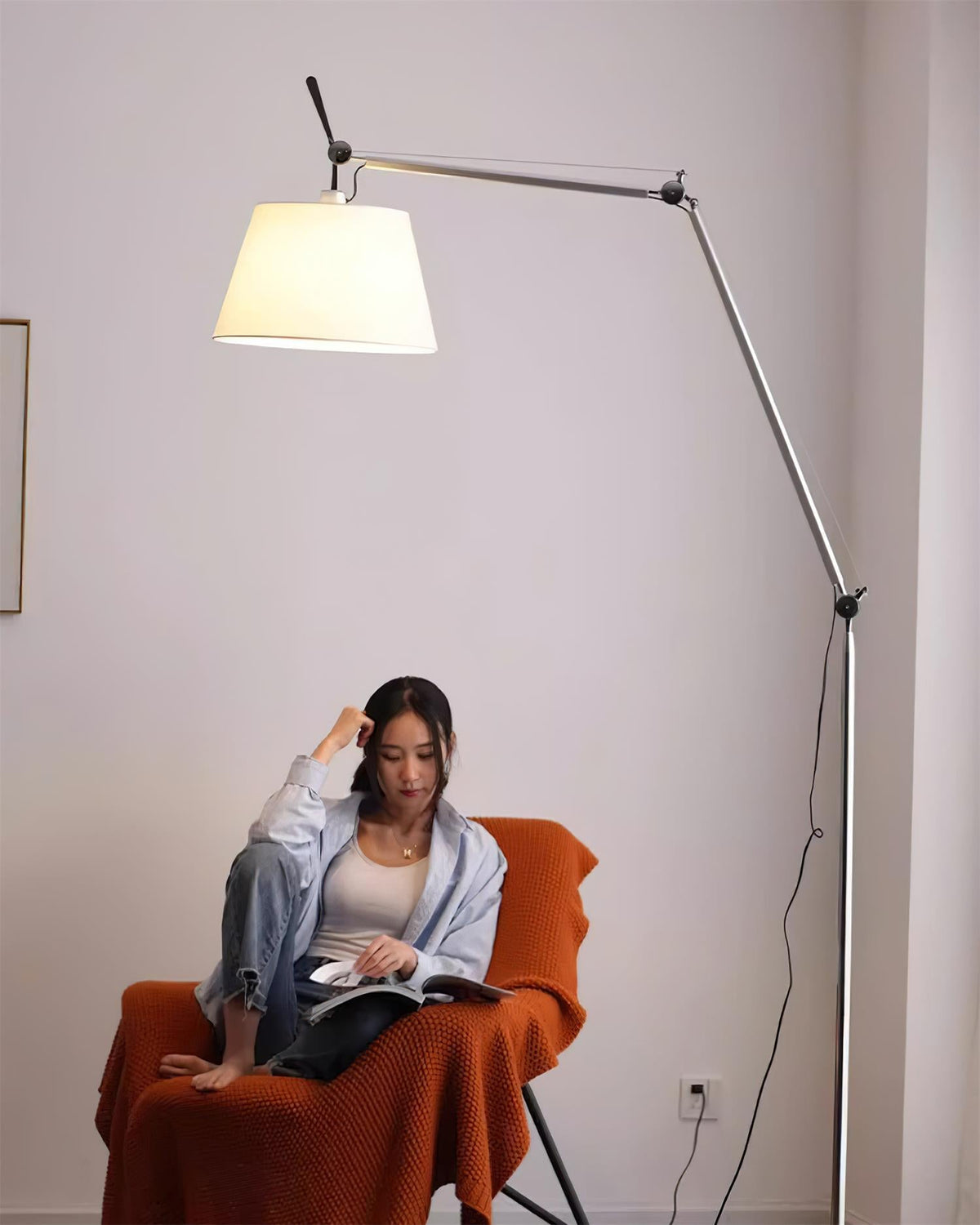 A woman reads in an orange armchair under the lit shade of this Clérachic floor lamp, its cord trailing down the pole.