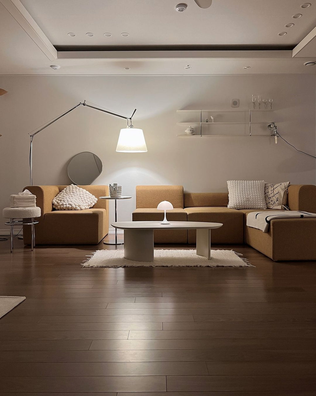 Clérachic floor lamp lit in a warm living room with a camel sofa, an oval white coffee table and a dark wood floor.
