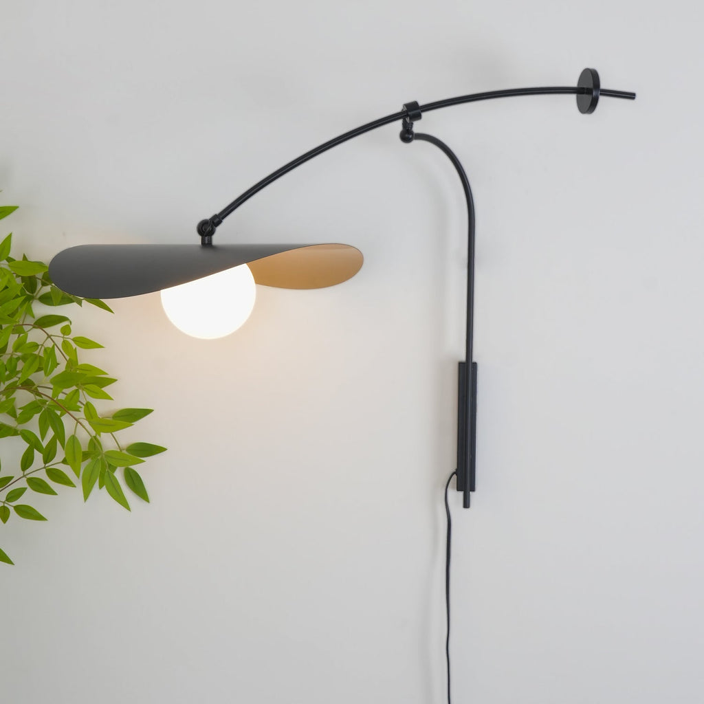 Close crop of this Clérachic wall light against a pale wall, a frosted white globe glowing under the black shade with green leaves beside it.