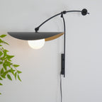 Angled view of a Clérachic wall light, its wide black shade tilted down over a white globe, with leafy branches at the left edge.