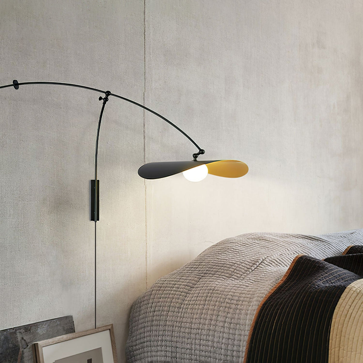 Above a grey knitted throw on a bed, this Clérachic wall light hangs its black arm and gold-lined shade against a textured wall.