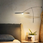 Low shade of this Clérachic wall light glowing over a dark headboard, with a plant and ceramic pots on a small round table.