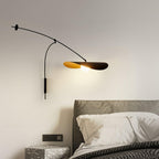 Bedside view of a Clérachic wall light over a grey headboard, with a patterned side table and a dark knit blanket.
