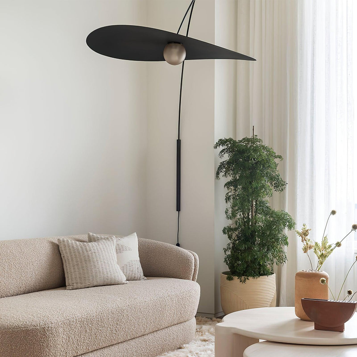 Raised high on its arm, this Clérachic wall light tilts a wide black shade above a boucle sofa and a potted tree.