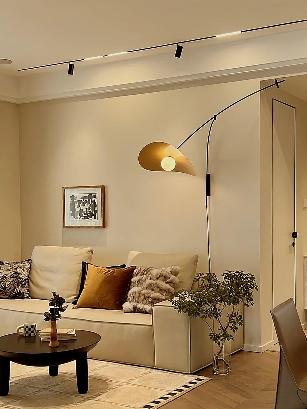 In a cream living room with track lights, this Clérachic wall light sends a warm glow from a gold-toned shade over a sofa.