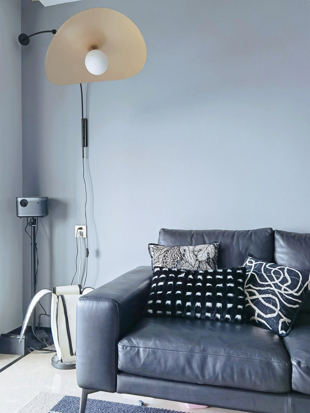 Customer-style photo of a Clérachic wall light on a blue-grey wall above a black leather sofa, the shade turned toward the camera.