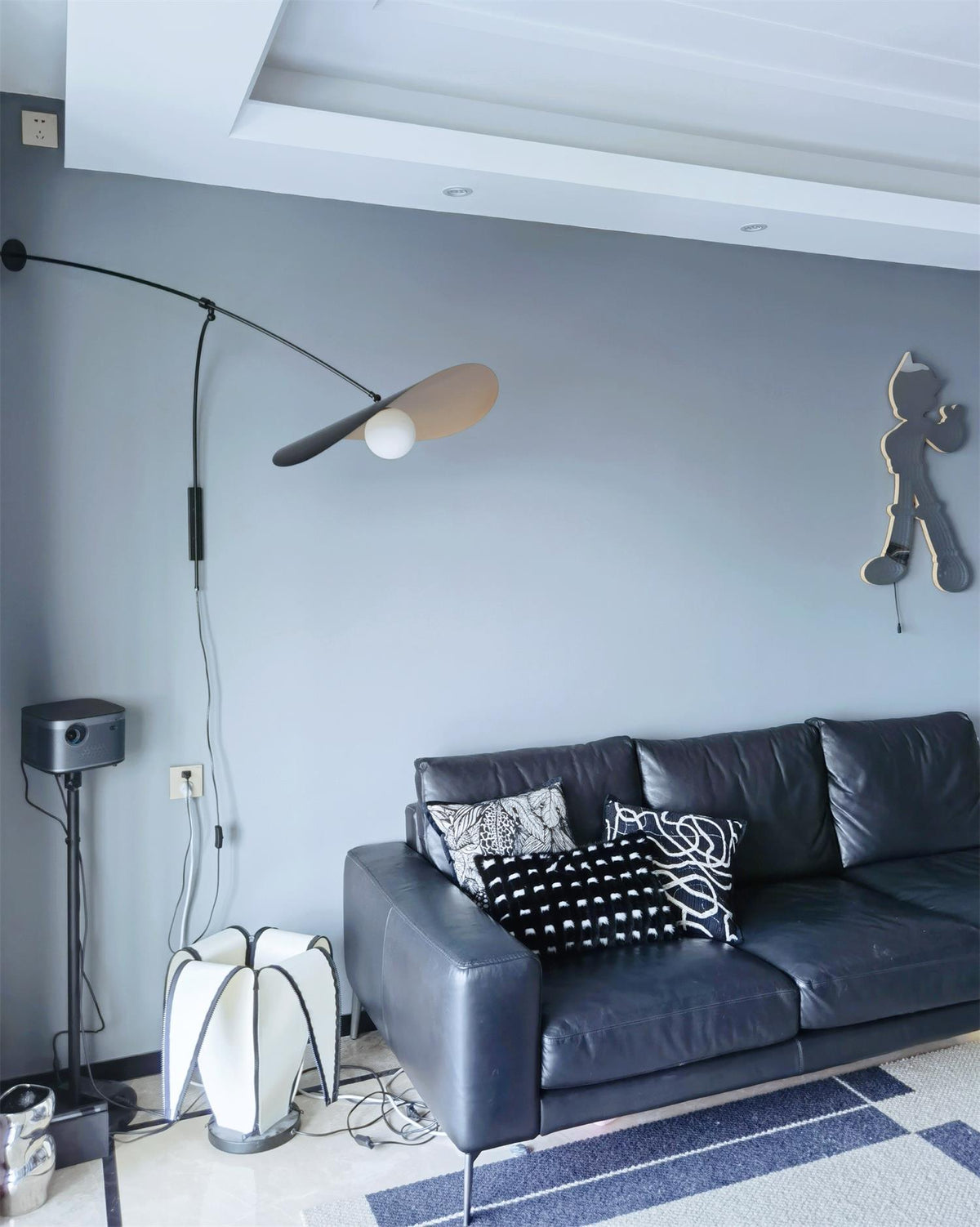 A Clérachic wall light hangs on a blue-grey wall above a dark leather sofa with patterned cushions, the shade angled sideways.