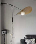Shade tilted up and lit, this Clérachic wall light sits on a grey wall with a projector and a patterned cushion below.