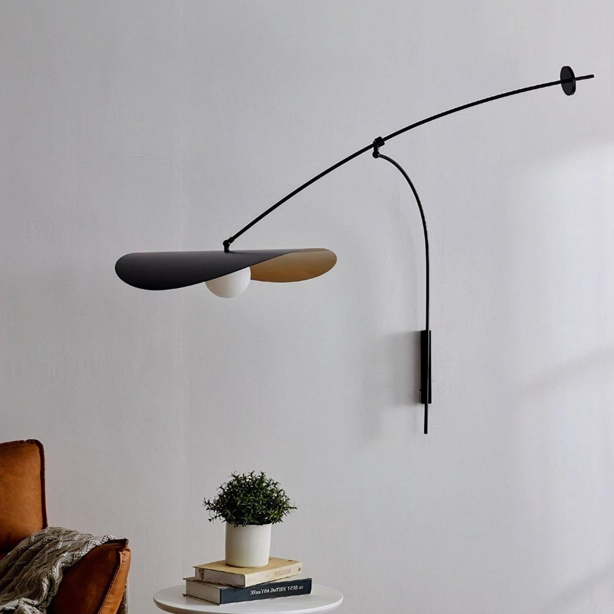 Close to a pale wall, this Clérachic wall light shows its black shade and a white globe, with a potted plant and books beneath.