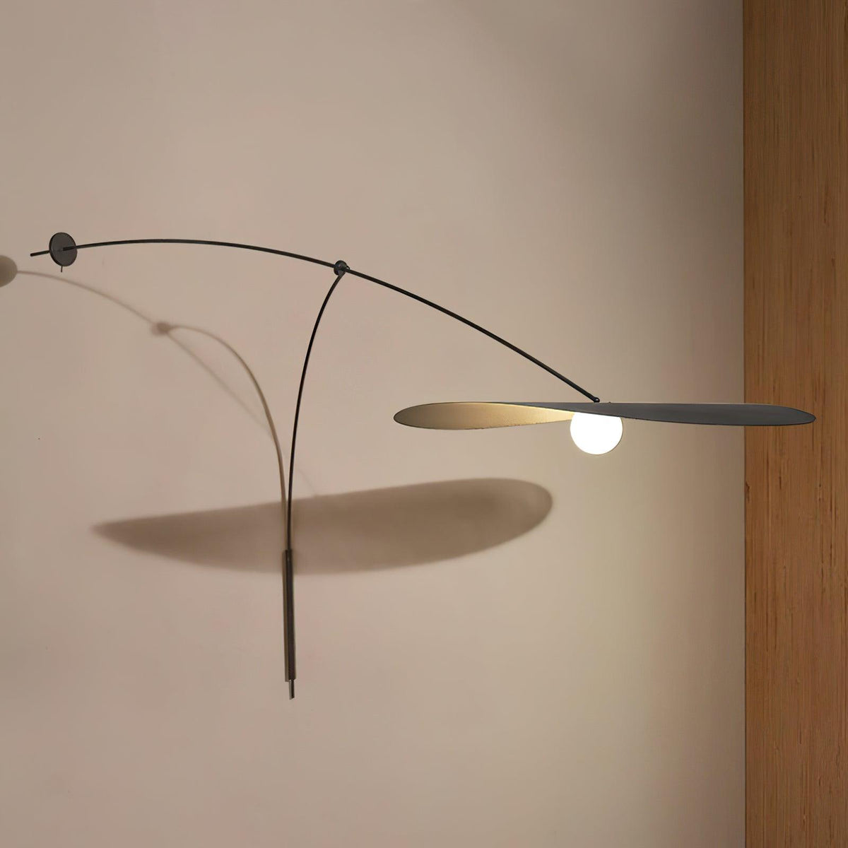 A Clérachic wall light with a long shadow on a warm beige wall, the black shade held flat beside a wooden panel.