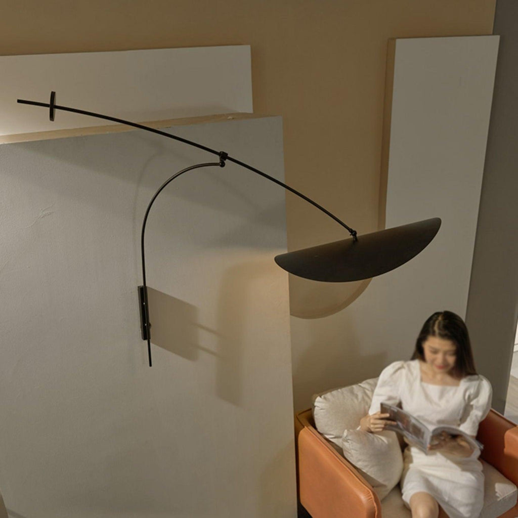 A reader on a sofa with a magazine sits below this Clérachic wall light, its black shade tilted against a pale wall.