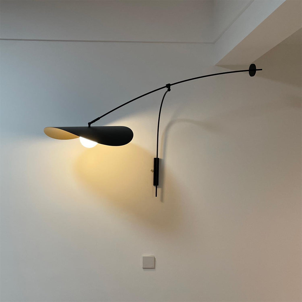 Everyday photo of a Clérachic wall light in a white room, its shade lit warm and tilted down toward a wall socket.