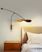 Narrow crop of this Clérachic wall light above a wooden nightstand and a rumpled bed, the gold-lined shade lit.