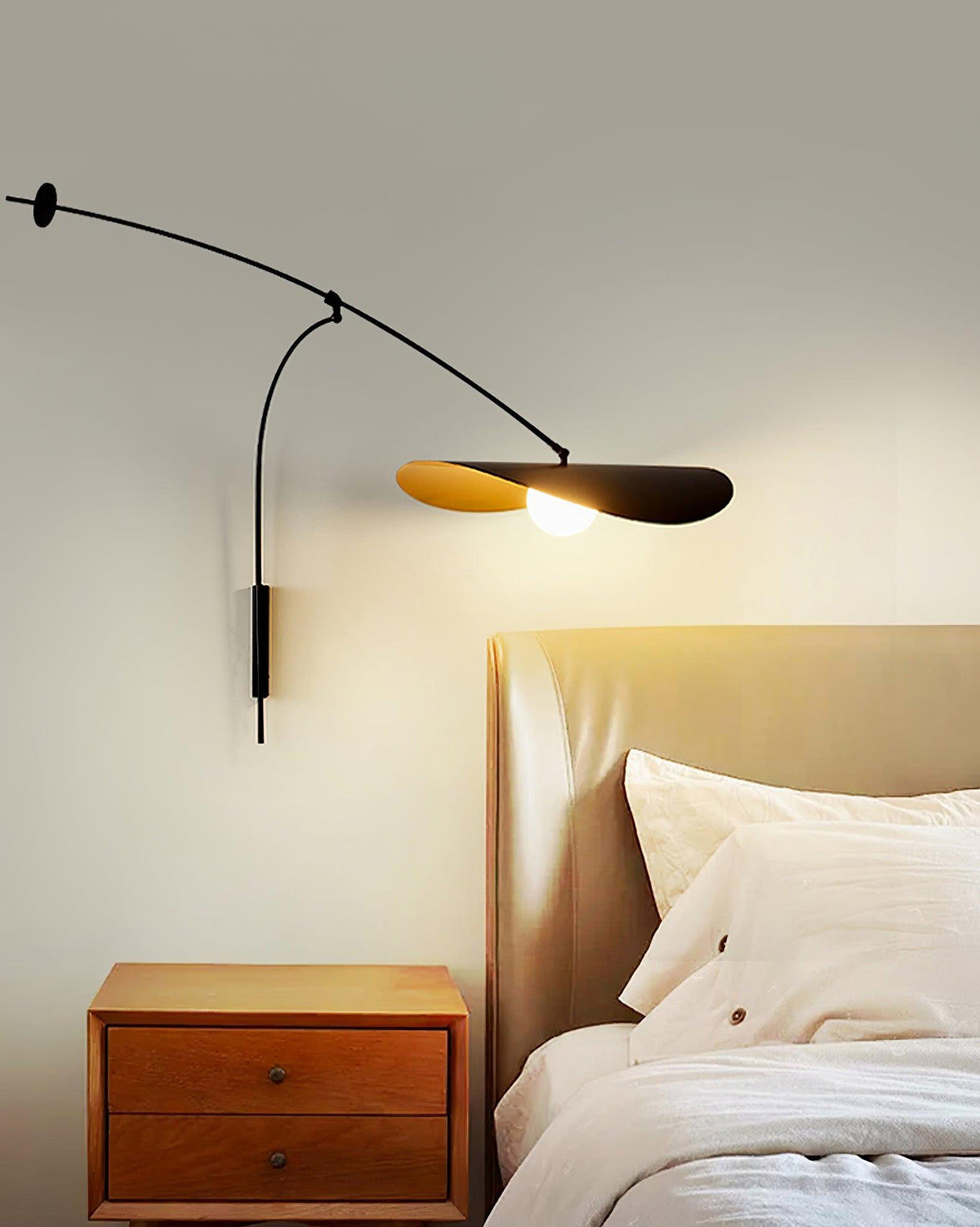 Narrow crop of this Clérachic wall light above a wooden nightstand and a rumpled bed, the gold-lined shade lit.