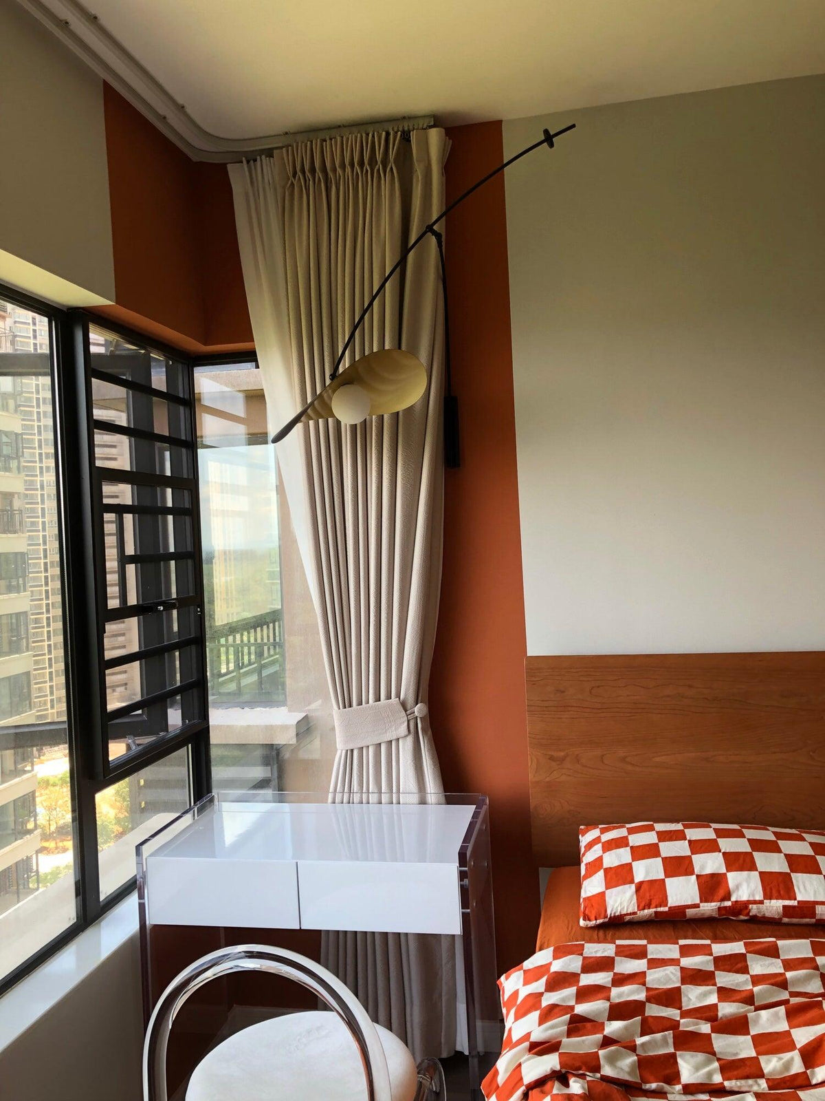 A Clérachic wall light on a bedroom wall beside a curtained window and a bed with orange checked bedding.