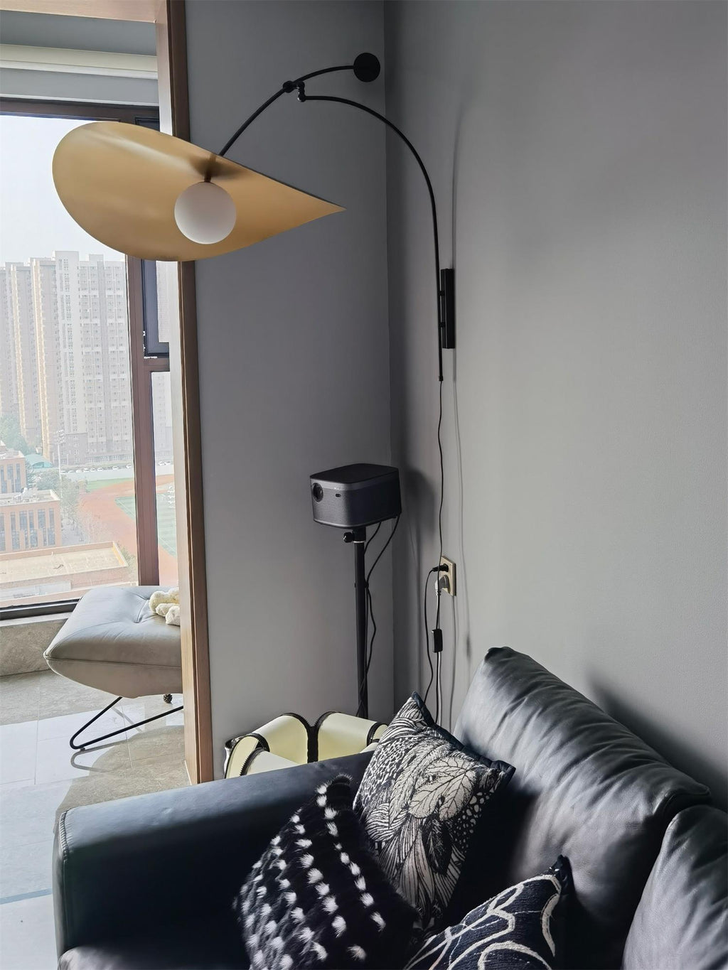 This Clérachic wall light's pale shade tilts forward by a tall window, above a black leather sofa and a projector.