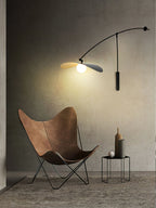Tall photo of a Clérachic wall light on a dark wall, glowing above a brown butterfly chair and a hexagonal side table.
