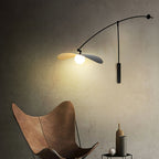Closer view of a Clérachic wall light lit warm above a leather butterfly chair, its black shade tilted slightly down.