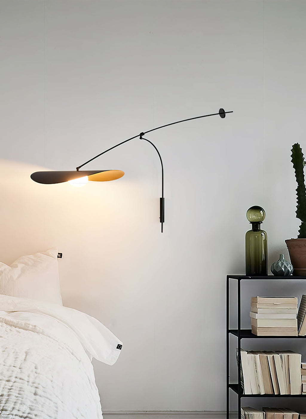 A Clérachic wall light lit beside a bed, with an open shelf of books and a green glass bottle nearby.