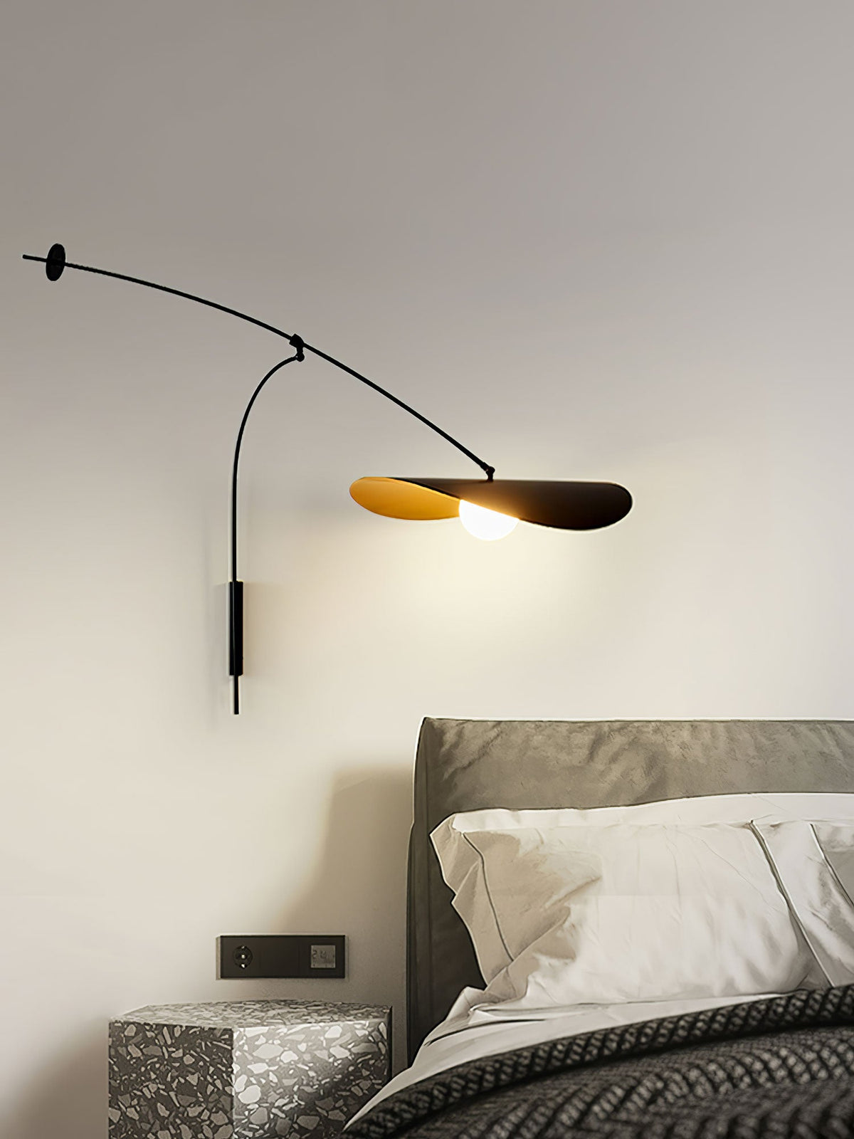 This Clérachic wall light glows over a grey headboard and white pillows, with a patterned side table at the bottom.