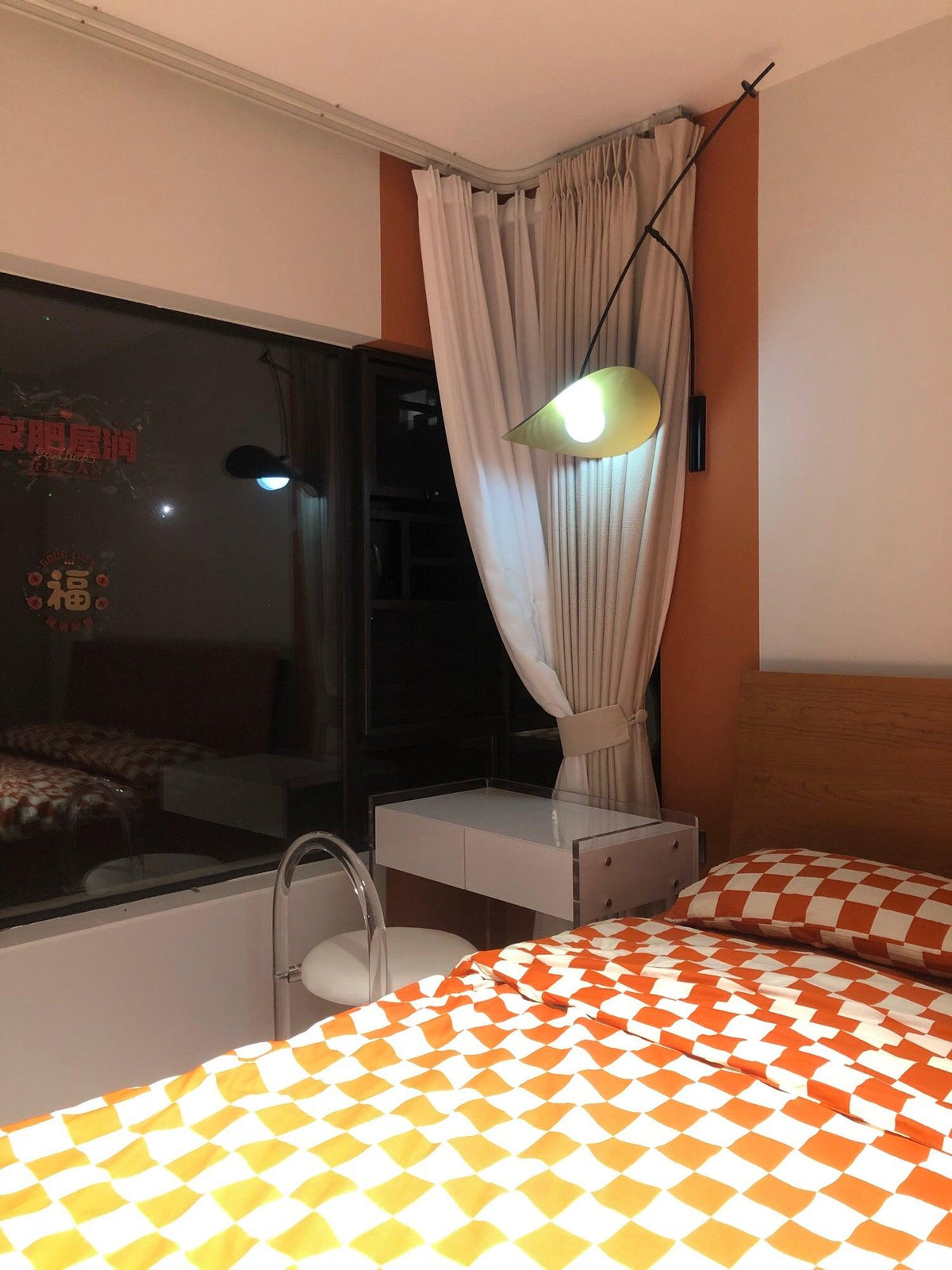At night in a bedroom with a curtained window, this Clérachic wall light shows a pale glowing shade above orange checked bedding.