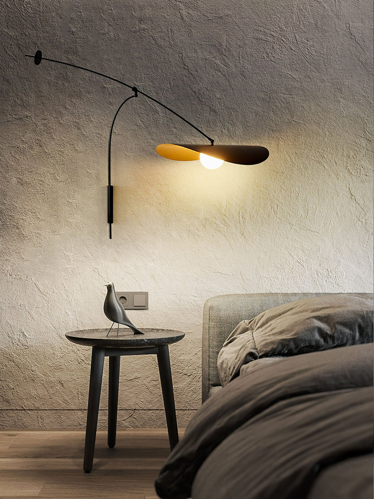 On rough grey plaster, this Clérachic wall light is lit above a round side table with a bird figure and a dark bed.