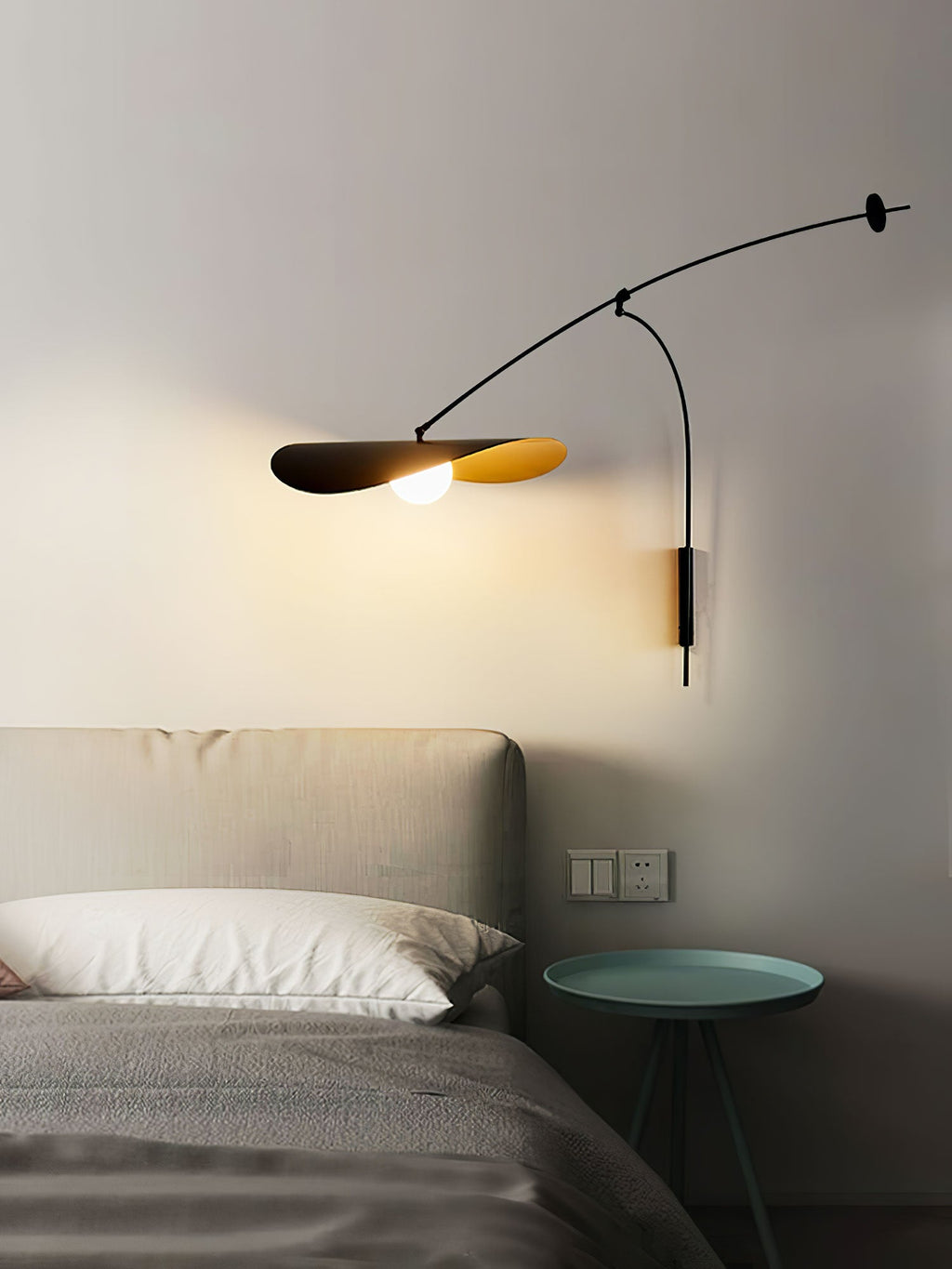 This Clérachic wall light lights a bedside corner, with a green side table, wall sockets and a grey bed below.