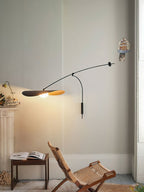 Over a wooden folding chair and a small table with an open book, this Clérachic wall light glows in a pale corner.