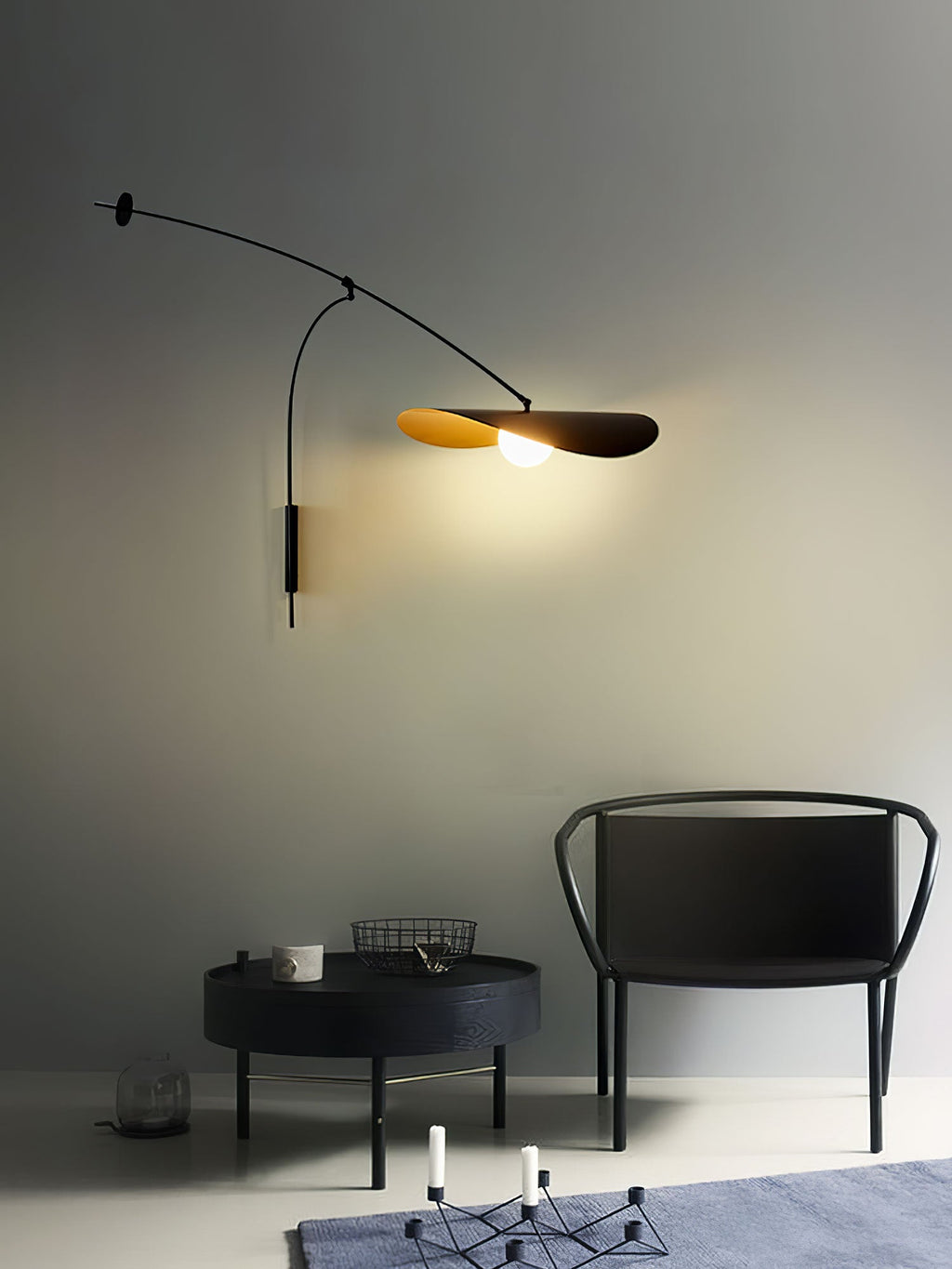 A Clérachic wall light with a gold-lined shade lights a grey wall above a black chair and a round dark coffee table.