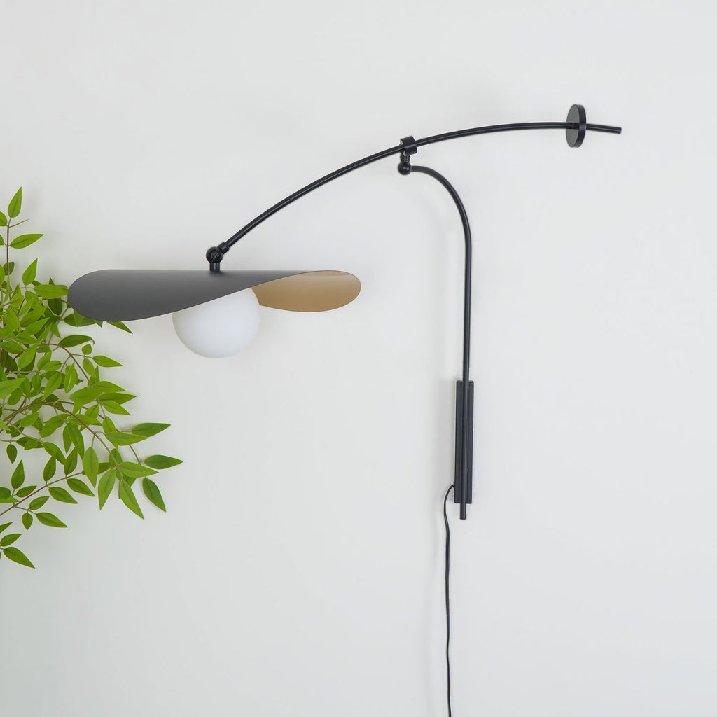 Leaf-lined view of a Clérachic wall light on a white wall, the black shade tilted over a white globe and a cord trailing down.