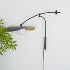 Leaf-lined view of a Clérachic wall light on a white wall, the black shade tilted over a white globe and a cord trailing down.