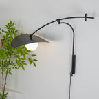 Lit white globe under the black shade of this Clérachic wall light, with a leafy plant beside the wall.