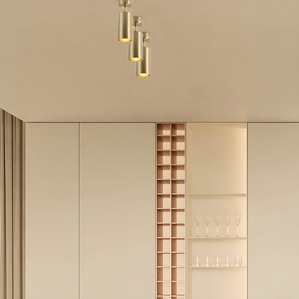 Three gold cylinder spotlights stepped down from a warm beige ceiling, with a shelving niche and glasses in the soft-lit room, from Clérachic.