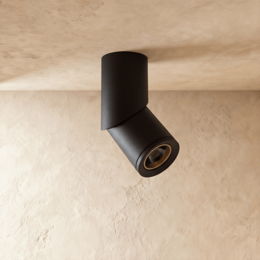 A black adjustable cylindrical downlight is angled from a plaster ceiling corner, its lens glowing warm, a Clérachic fixture close up.